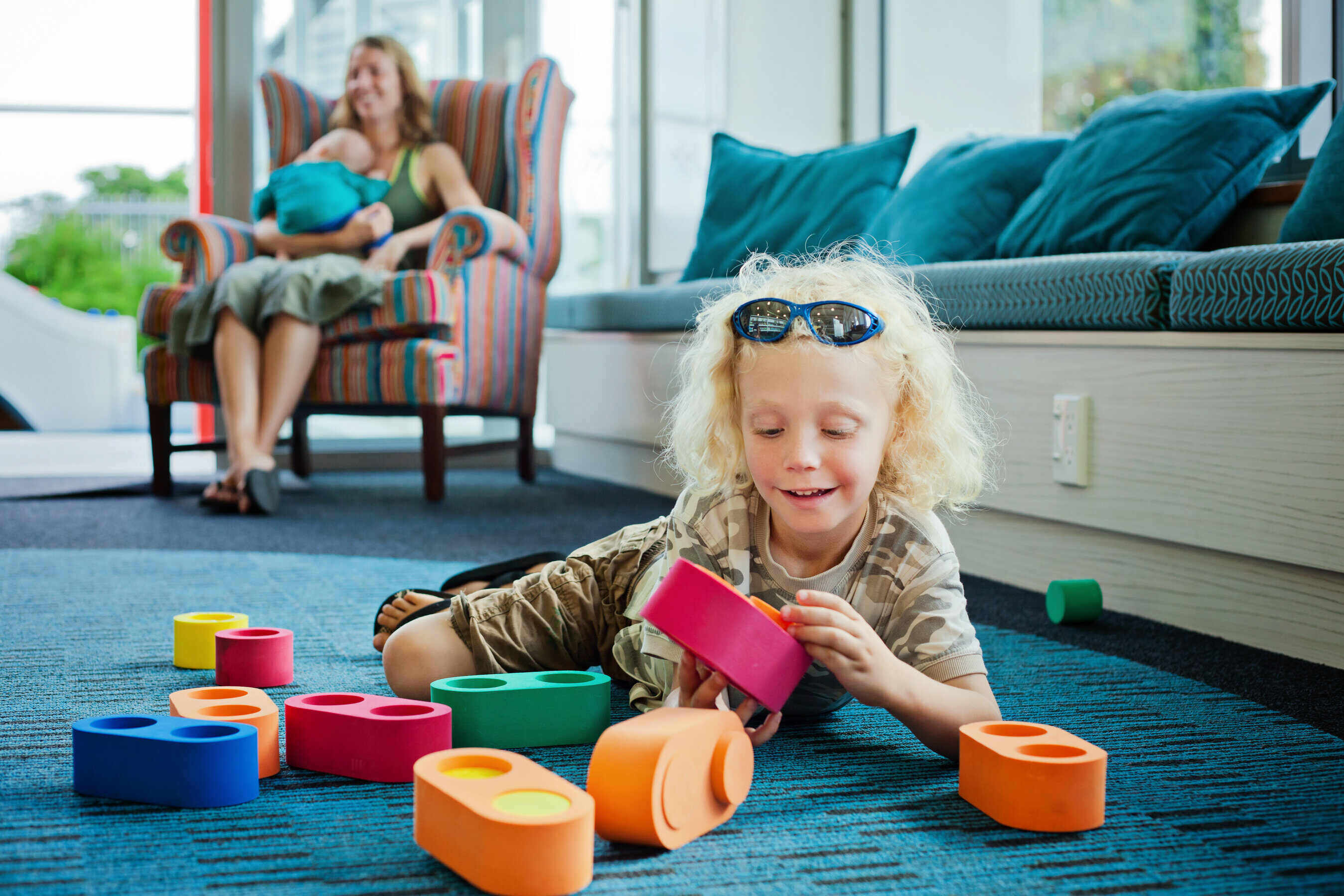 Image of a child with curly blonde hair playing with blocks while his mother has a child on her chest