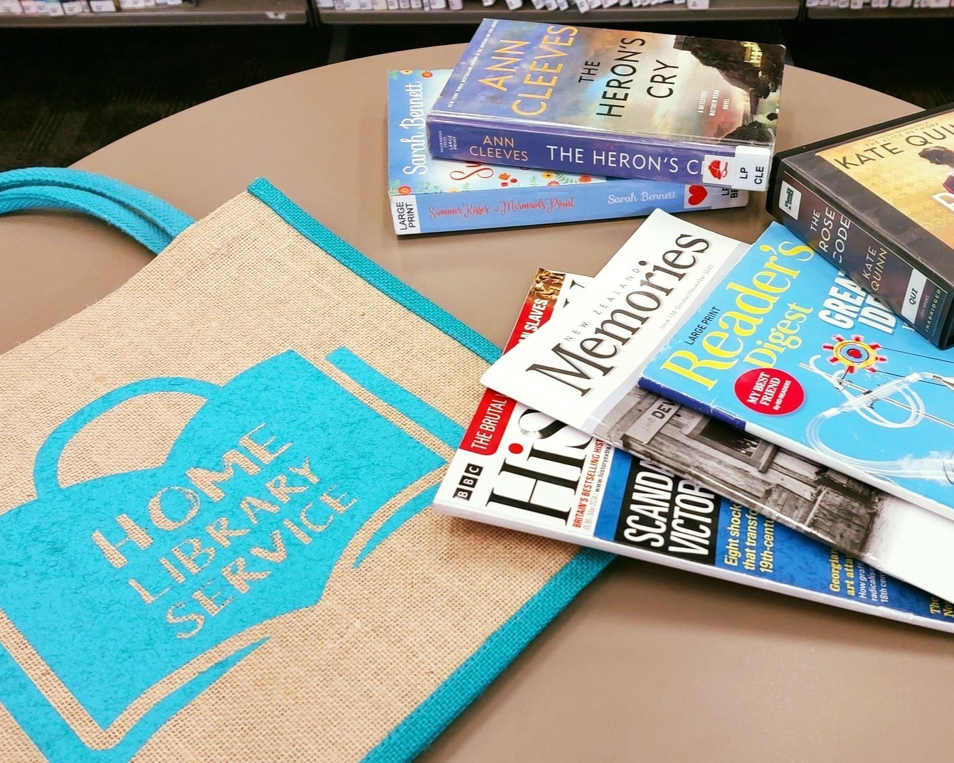 Image shows a carry bag with 'Home Library Service' on it. It also shows three magazines, two large print books, and one audio book as examples as some of the items you might borrow from a home library service.